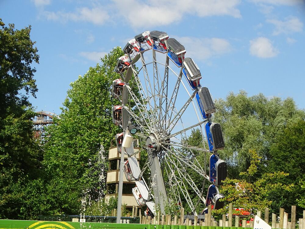 Grote wielvormige kermisattractie met gekleurde gondels boven groene bomen tegen een blauwe lucht
